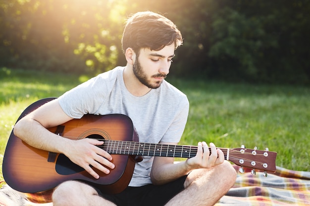 Retrato horizontal del hombre barbudo de moda sentado con las piernas cruzadas tocando la guitarra