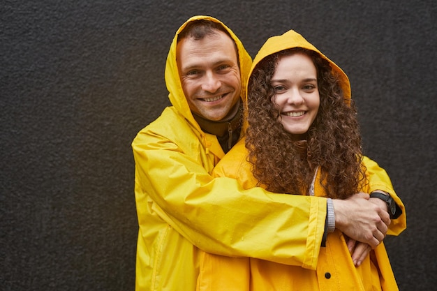 Retrato horizontal de un hombre y una mujer caucásicos enamorados usando impermeables amarillos abrazándose y mirando la cámara