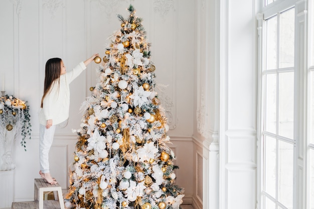 El retrato horizontal de una niña pequeña se para en la silla, decora el árbol de Navidad, trata de mostrarse mejor, está en casa, disfruta de un ambiente tranquilo y doméstico, quiere satisfacer a los padres, trabaja duro