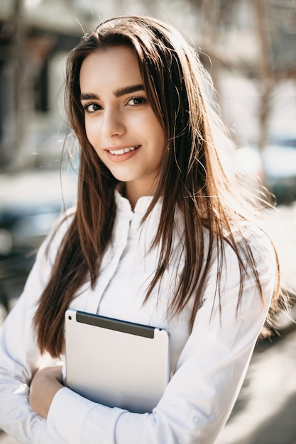 Retrato de una increíble mujer de pelo largo mirando a la cámara confiada mientras sostiene una tableta vestida con camisa blanca afuera en la calle.