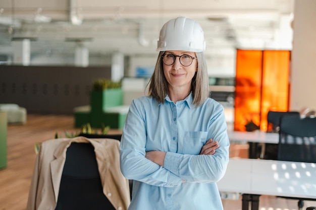 Retrato de una ingeniera arquitecta seria y segura de sí misma sosteniendo los brazos cruzados usando casco mirando la cámara de pie en una oficina moderna Concepto de negocio exitoso