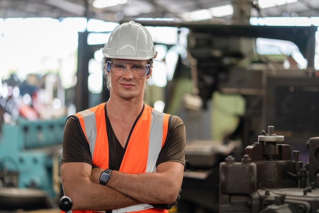 Foto retrato de ingeniero masculino en chaleco de seguridad con casco sonriendo y cruzando el brazo en fábrica