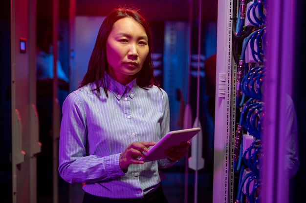 Retrato de un ingeniero de redes asiático serio que soluciona problemas mientras trabaja con una supercomputadora en la sala de servidores, luz de neón oscura