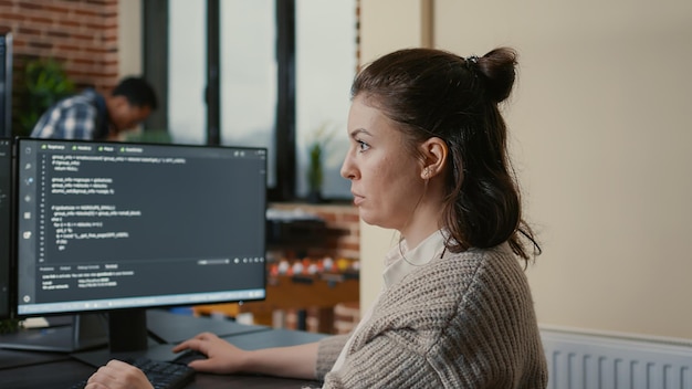 Retrato de un ingeniero de software enfocado que escribe código mirando varias pantallas de computadora que muestran un algoritmo de lenguaje de programación. El programador codifica mientras sus colegas desarrollan aplicaciones de trabajo en equipo.