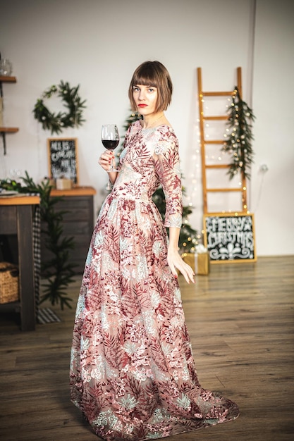 Retrato interior de menina confiante em um vestido longo, se passando perto da árvore de Natal com um copo de vinho. Foco suave. Foco seletivo.