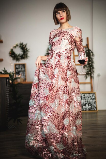 Retrato interior de menina confiante em um vestido longo, se passando perto da árvore de Natal com um copo de vinho. Foco suave. Foco seletivo.