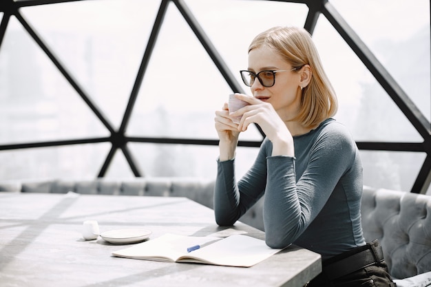 Retrato interior de uma jovem mulher de negócios sentada em um café e bebendo um café. Rapariga loira com óculos escuros e jaqueta