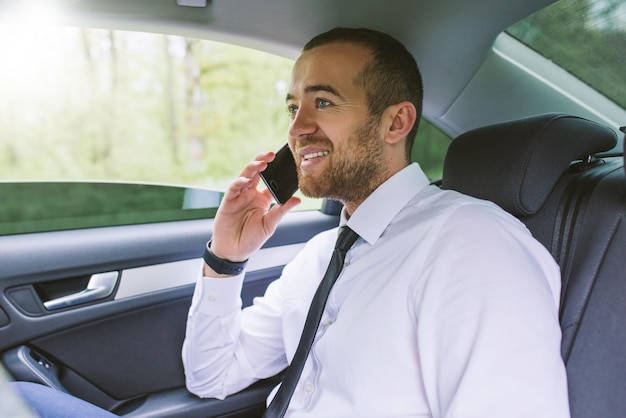 Retrato interior de un hombre de negocios feliz hablando por teléfono móvil sentado en un asiento trasero del automóvil Hombre de negocios de Handosme usando un teléfono inteligente para hablar sobre noticias y éxito Concepto de tecnología y negocios