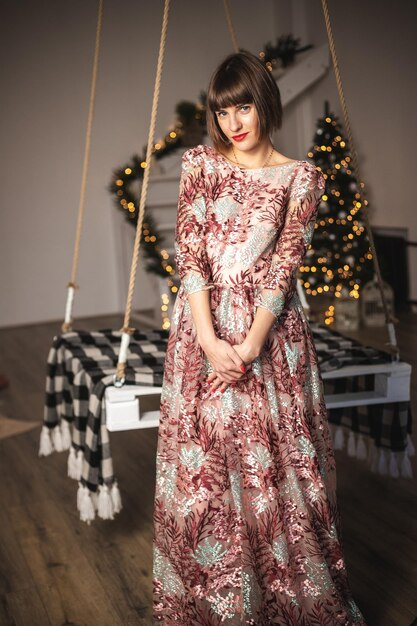 Retrato de interior de niña confiada en un vestido largo posando cerca del árbol de Navidad en el columpio. Enfoque suave. Enfoque selectivo.