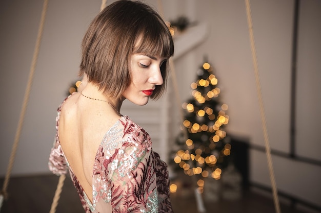 Retrato de interior de niña confiada en un vestido largo posando cerca del árbol de Navidad. Enfoque suave. Enfoque selectivo.