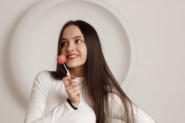 Foto retrato interno de uma jovem morena sorridente positiva sobre fundo branco, segurando um bolo de sobremesa doce nas mãos