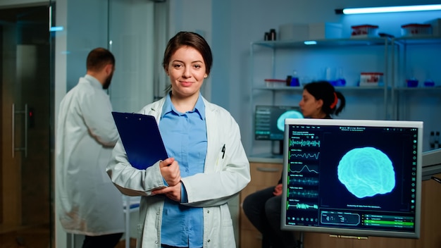 Retrato del investigador neurólogo médico mirando a la cámara sonriendo sosteniendo clipdoard mientras un compañero de trabajo discutiendo con el paciente sobre la atención de la salud del cerebro