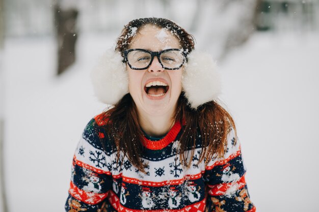 Retrato de invierno cerca al aire libre de joven hermosa con orejeras, suéter posando en el parque cubierto de nieve. Mujer mirando y sonriendo