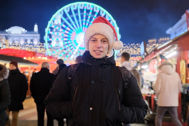 Retrato de invierno de feliz joven adolescente en el mercado de Navidad. Luces, guirnaldas brillantes, fondo de noria.