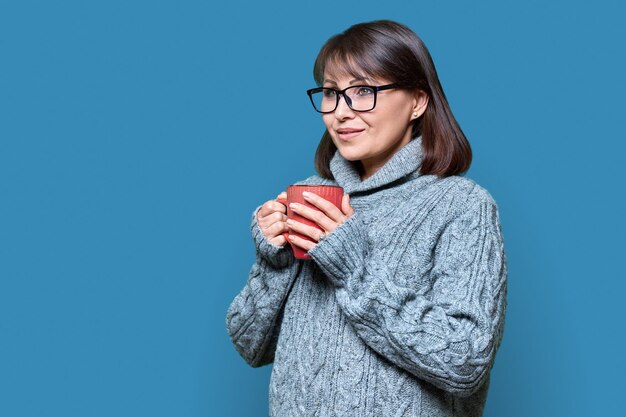 Retrato de invierno de una mujer feliz y madura en suéter con taza en fondo azul de estudio sonriente vista de perfil femenino belleza de invierno estilo de vida acogedor concepto de Navidad de Año Nuevo