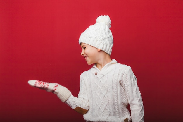 Retrato de invierno de un niño feliz y alegre con un sombrero de punto y un suéter divirtiéndose en un fondo rojo, el niño mira sus palmas