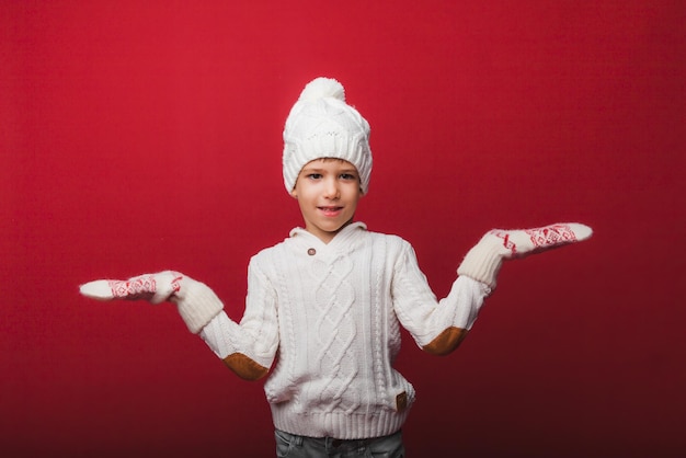 Retrato de invierno de un niño feliz y alegre con un sombrero de punto y un suéter divirtiéndose en un fondo rojo, el niño mira sus palmas