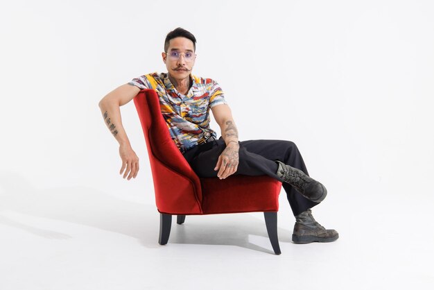 Retrato isolado cortado em estúdio tiro asiático vintage elegante bigode pescoço braços mãos tatuagens modelo de moda masculino em casual roupa clássica botas de couro sentado em cadeira vermelha fundo branco