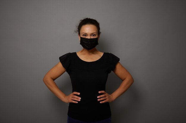 Retrato isolado em fundo cinza de uma mulher em máscara protetora médica preta, olhando para a câmera com as mãos na cintura. Copie o espaço para anúncio. Conceito de Black Friday