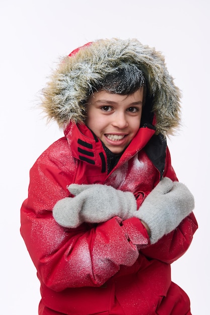 Foto retrato isolado no fundo branco de um belo menino de idade elementar caucasiano, vestido com uma parka vermelha brilhante e um capuz coberto de neve, olhando para a câmera enquanto se abraça, tremendo de frio