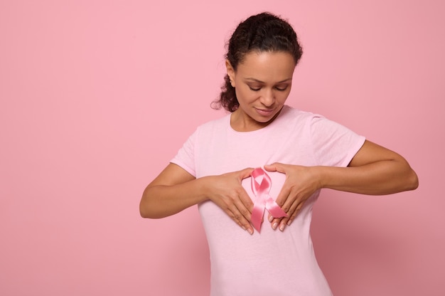 Foto retrato isolado no fundo colorido de uma mulher afro-americana em uma camiseta rosa, colocando as mãos no peito em forma de coração com uma fita de cetim rosa no centro. dia mundial da conscientização do câncer.