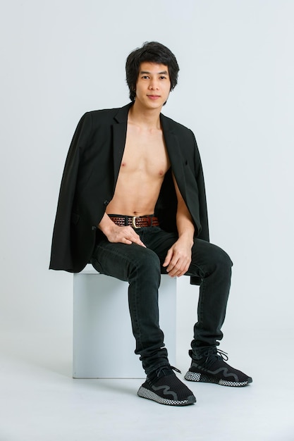 Retrato isolado tiro de estúdio asiático jovem bonito confiante magro magro sem camisa saudável atlético moda adolescente modelo masculino em terno blazer jeans e tênis sentado sorrindo posando em fundo branco