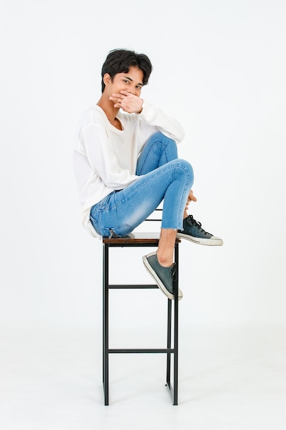 Retrato isolado tiro de estúdio asiático jovem LGBT gay feliz bonito bissexual homossexual modelo masculino em roupa casual sentado de pernas cruzadas na cadeira alta sorrindo olhar para a câmera no fundo branco