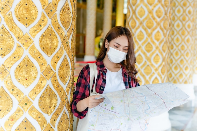 Retrato jovem asiático mochileiro feminino na máscara em pé e segurando o mapa de papel na mão no belo templo tailandês, ela procurando direção