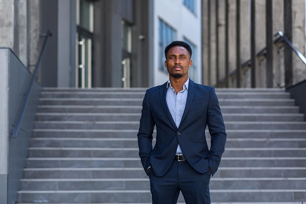 Retrato jovem confiante bem sucedido empresário afro-americano, olhando para a câmera homem de negócios sério em pé no meio urbano de um edifício de escritórios moderno ao ar livre. traje formal masculino, braços cruzados