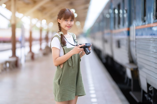 Retrato Jovem mulher asiática sorrindo turista Garota viajante andando e segurando a câmera espera viagem de trem é tirada na plataforma ferroviária Tailândia verão relaxar férias Conceito