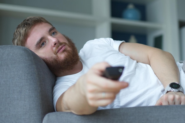 Retrato de un joven aburrido con ropa informal sentado y acostado en un sofá en la sala de estar y mirando la televisión