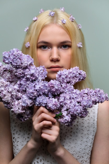 Retrato de joven adolescente rubia con flores lilas violetas en el cabello y las manos, fondo de estudio verde pastel. Piel sana y maquillaje natural para adolescentes