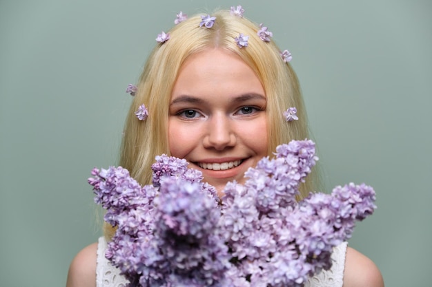 Retrato de joven adolescente rubia con flores lilas violetas en el cabello y las manos, fondo de estudio verde pastel. Piel sana y maquillaje natural para adolescentes