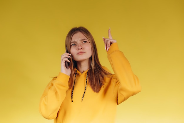 Retrato de una joven adolescente sosteniendo un teléfono una chica hablando por teléfono con una sudadera con capucha amarilla casual en un tono con el fondo