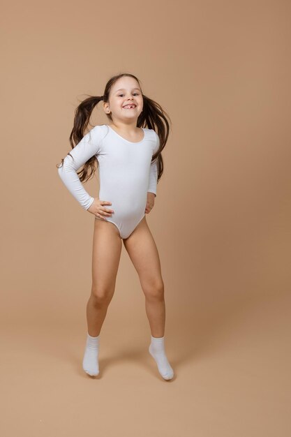 Foto retrato de joven adorable niña sonriente feliz con cabello largo y oscuro en traje de baño de entrenamiento blanco y calcetines saltando sobre fondo marrón posando divirtiéndose