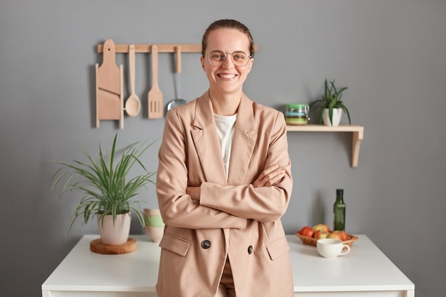 Retrato de una joven adulta caucásica sonriente mujer de negocios vestida con traje beige y anteojos de pie en la cocina mirando a la cámara con una sonrisa mantiene los brazos cruzados