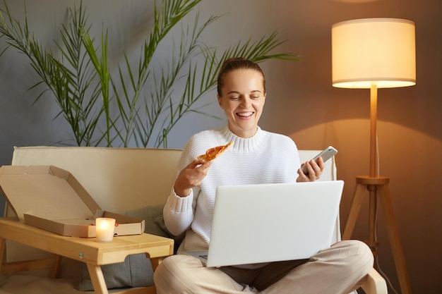 Retrato de una joven adulta caucásica sonriente con suéter blanco sentada en un sofá y trabajando en una laptop sosteniendo en las manos una rebanada de pizza y un teléfono móvil