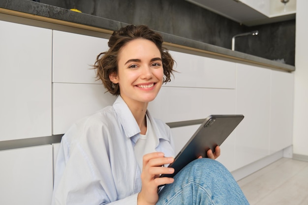 Retrato de una joven adulta que trabaja en su tableta y se sienta con un cuaderno en el suelo de la cocina en casa