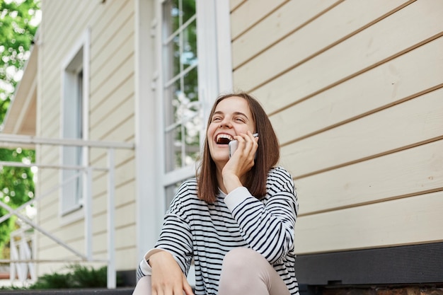 Retrato de una joven adulta riendo hablando con un teléfono móvil al aire libre cerca del edificio comunicándose con amigos pasando un buen rato y una conversación agradable