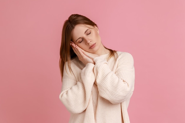 Retrato de una joven adulta rubia cansada y exhausta durmiendo acostada en las palmas de las manos manteniendo los ojos cerrados usando suéter blanco Foto de estudio interior aislada en fondo rosa