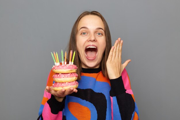 Retrato de una joven adulta sorprendida y sorprendida con un suéter cálido sosteniendo un donut con velas aisladas sobre un fondo gris con los brazos levantados gritando de felicidad