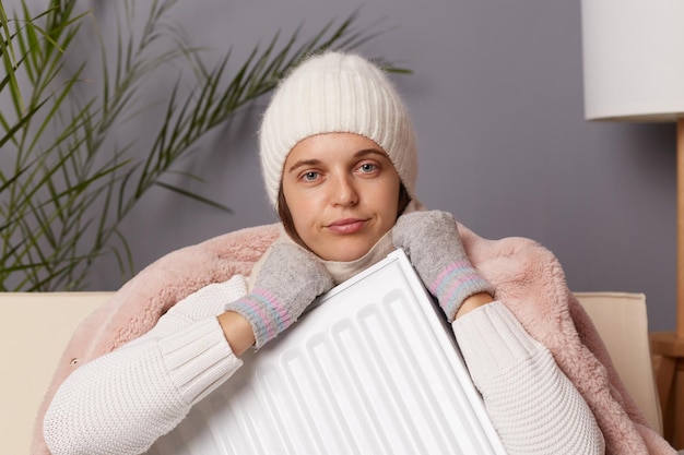 Retrato de una joven adulta triste y decepcionada con abrigo y sombrero sentada en una sala de estar fría con calentador en las manos mirando la cámara con expresión molesta y labios fruncidos