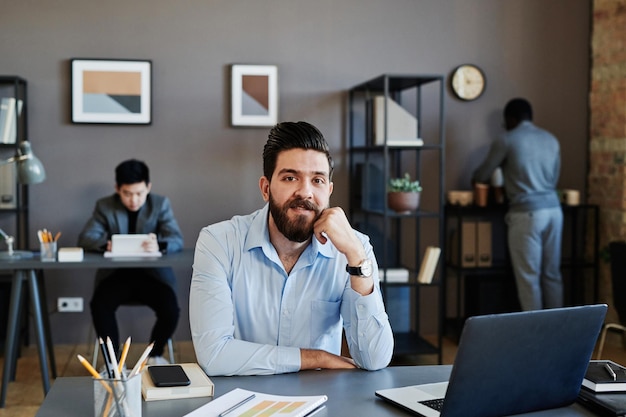 Foto retrato de un joven adulto con barba en la cara vestido con una camisa azul sentado en el escritorio en la oficina mirando a la cámara