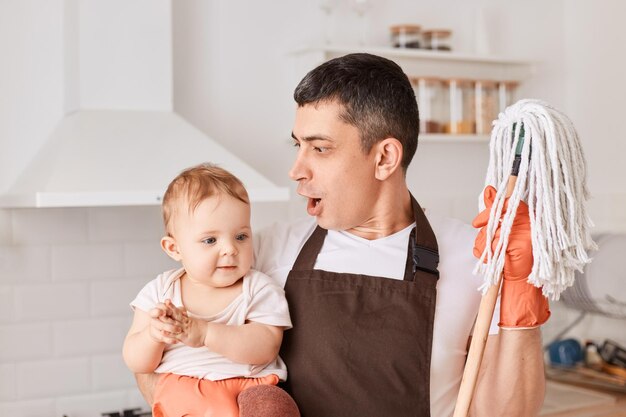 Retrato de un joven adulto conmocionado y una niña sosteniendo trapeadores limpiando en la cocina usando un delantal marrón mirando a un niño con una expresión sorprendida y asombrada haciendo tareas domésticas