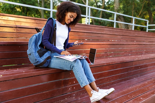 Retrato de una joven africana feliz con mochila mediante teléfono móvil mientras descansa en el parque, leyendo una revista