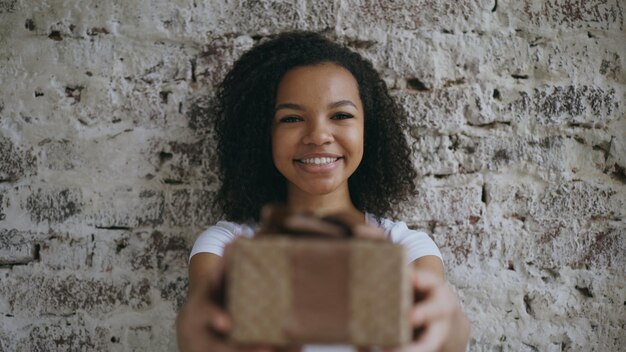 Retrato de una joven africana feliz que presenta una caja de regalo y sonríe a la cámara