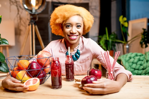 Retrato de una joven africana con frutas rojas frescas y batidos sentado en el interior en el acogedor interior de la casa