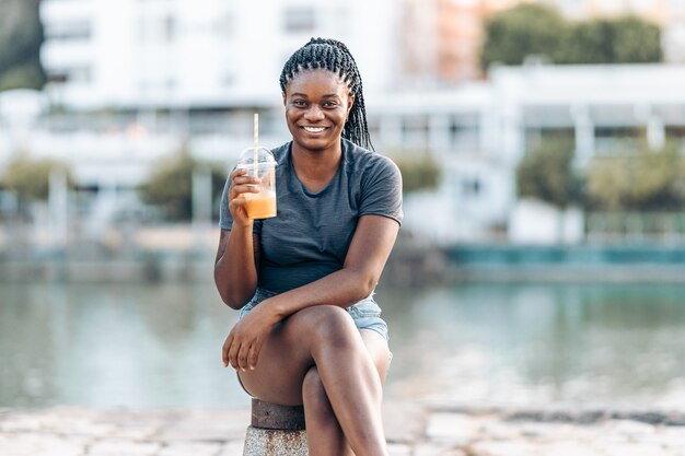 Retrato de una joven africana sentada mientras sostiene un batido y sonriendo a la cámara al aire libre