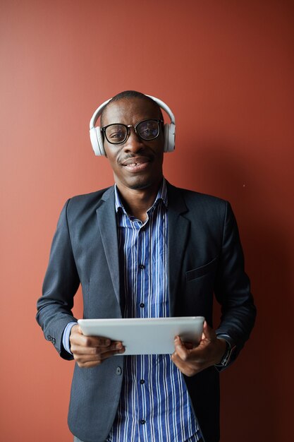 Retrato de joven africano en anteojos y auriculares inalámbricos sonriendo a la cámara mientras usa tablet pc contra el fondo marrón