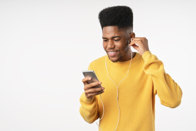 Retrato de un joven africano casual sonriente con teléfono y auriculares parados sobre fondo blanco